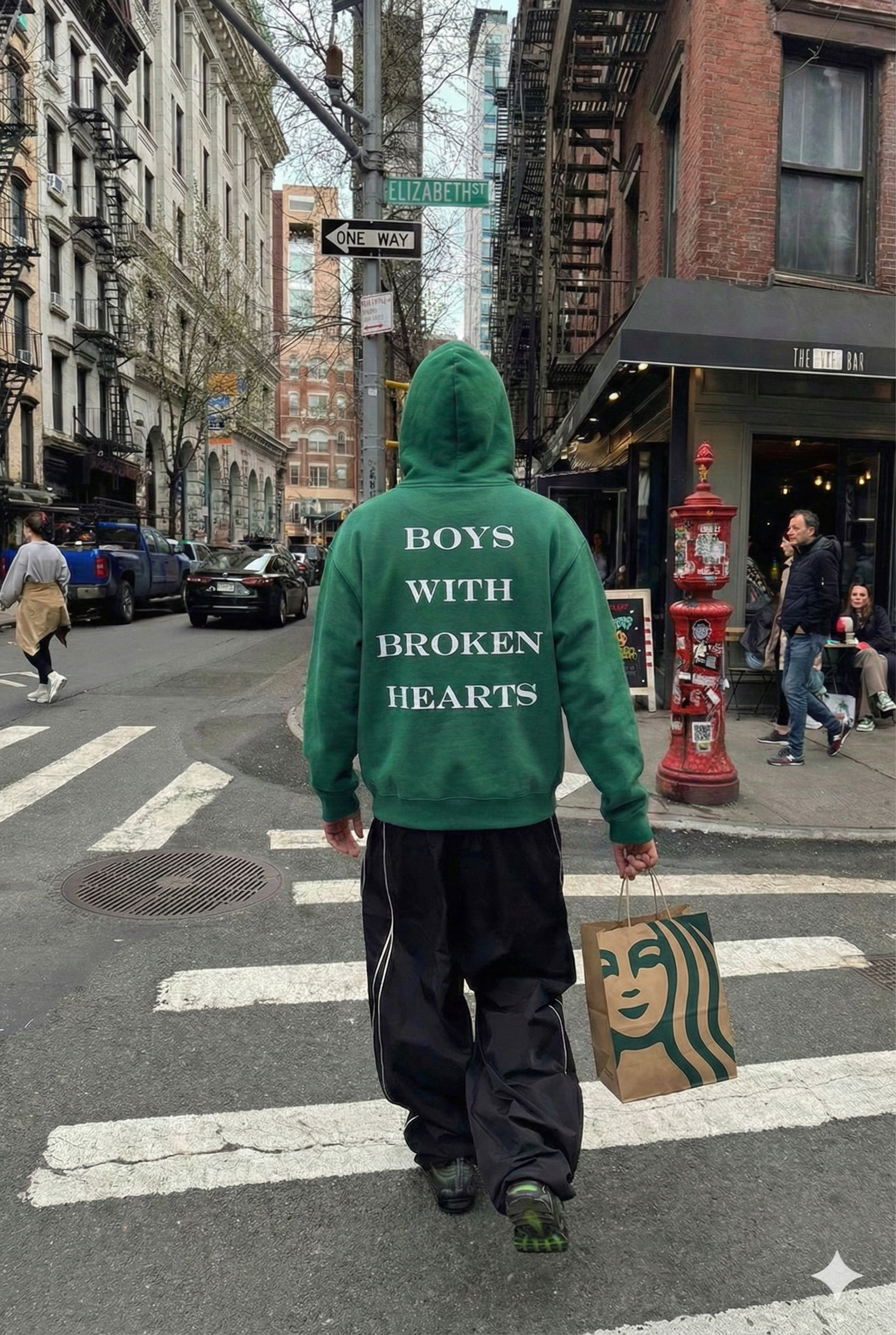 Person wearing a boys with broken hearts green hoodie with text on the back, walking on a city street.